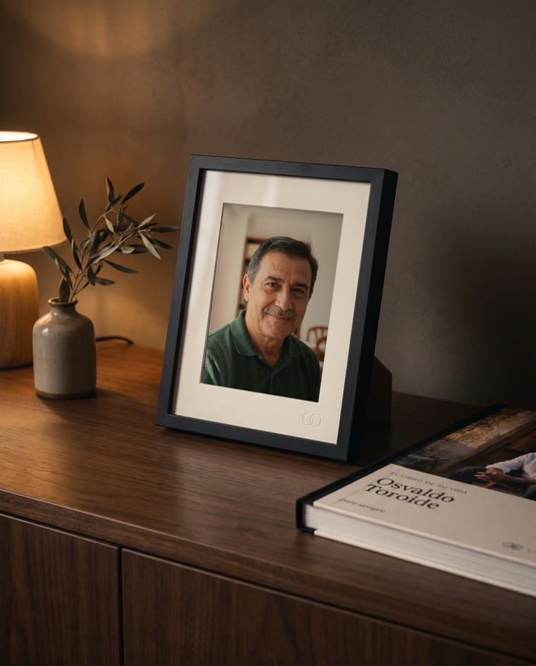 Un marco negro chico con una foto, sobre un aparador junto a un libro y una lámpara prendida