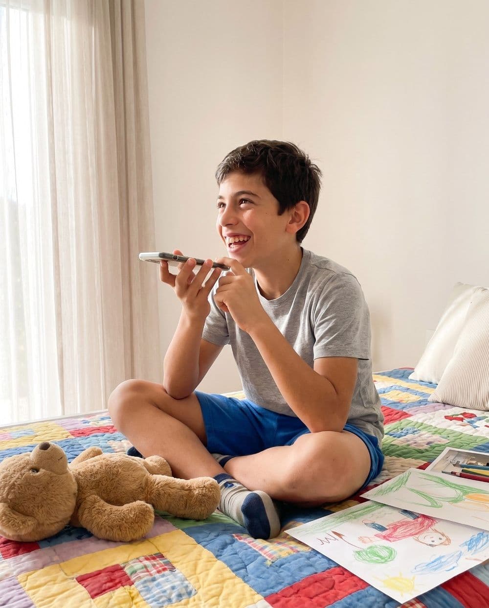 Un chico sentado en su cama graba un audio en el celular, entre dibujos y un oso de peluche
